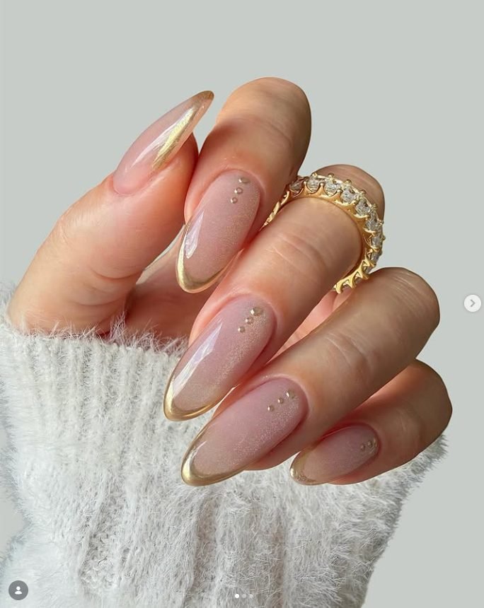 Long almond nude nails with a soft cat-eye sparkle, gold chrome French tips and tiny gold studs