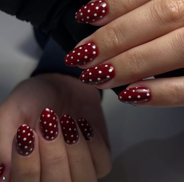 Glossy cranberry red almond nails covered in small white polka dots