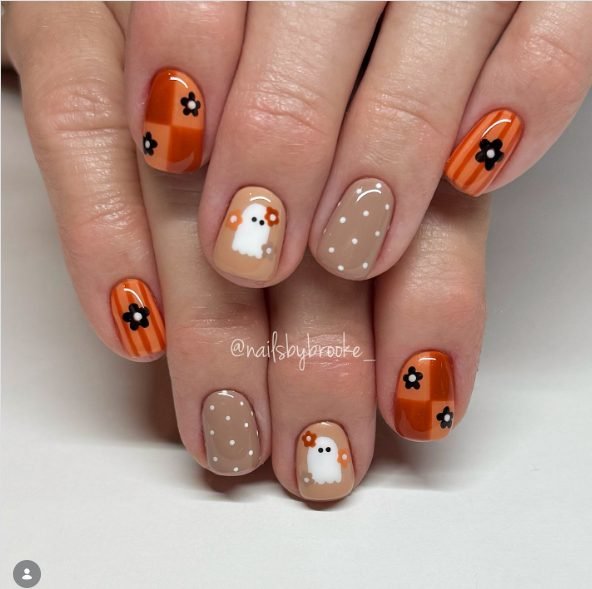 Short round nails with orange checks and stripes, black flowers, tan polka dots and white ghosts holding flowers