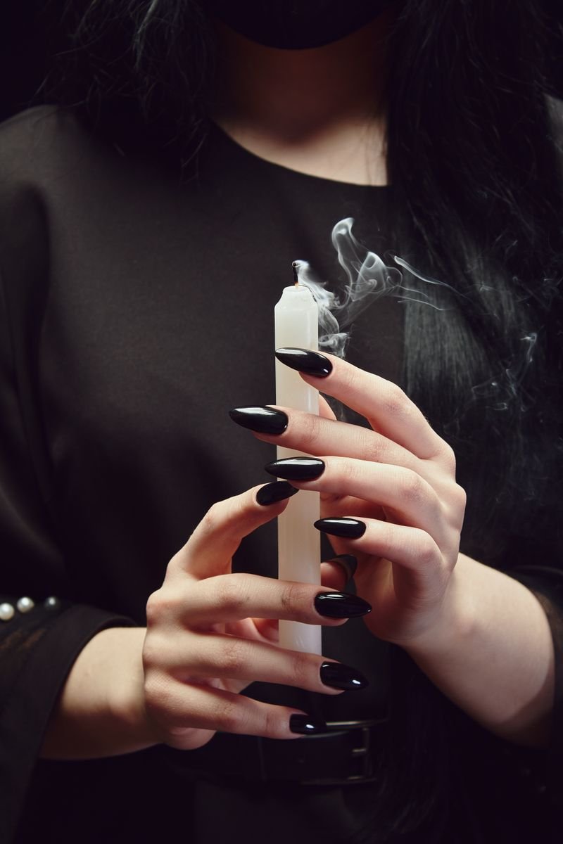 Close-up of hands with black polished nails holding a white candle, creating a mysterious ambiance.